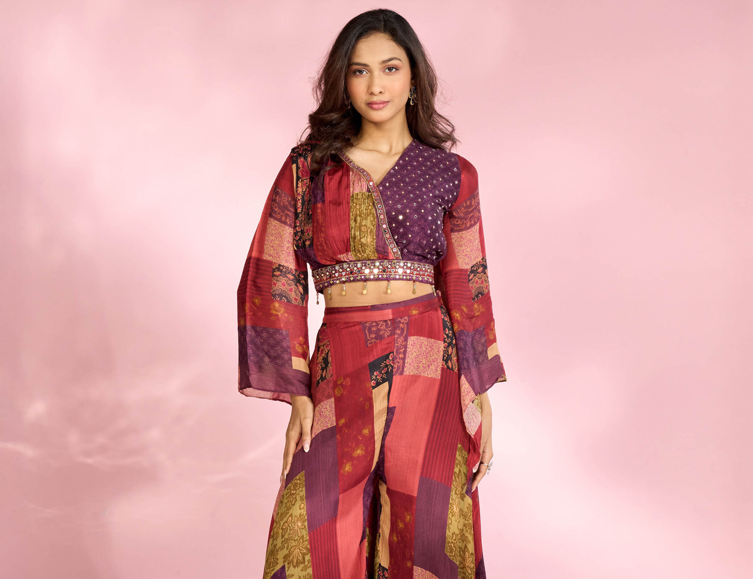 Young woman in a stylish maroon and multicolor patchwork co-ord set with flared sleeves and embroidered waist detail, posing confidently against a soft pink background for Navratri festive fashion.